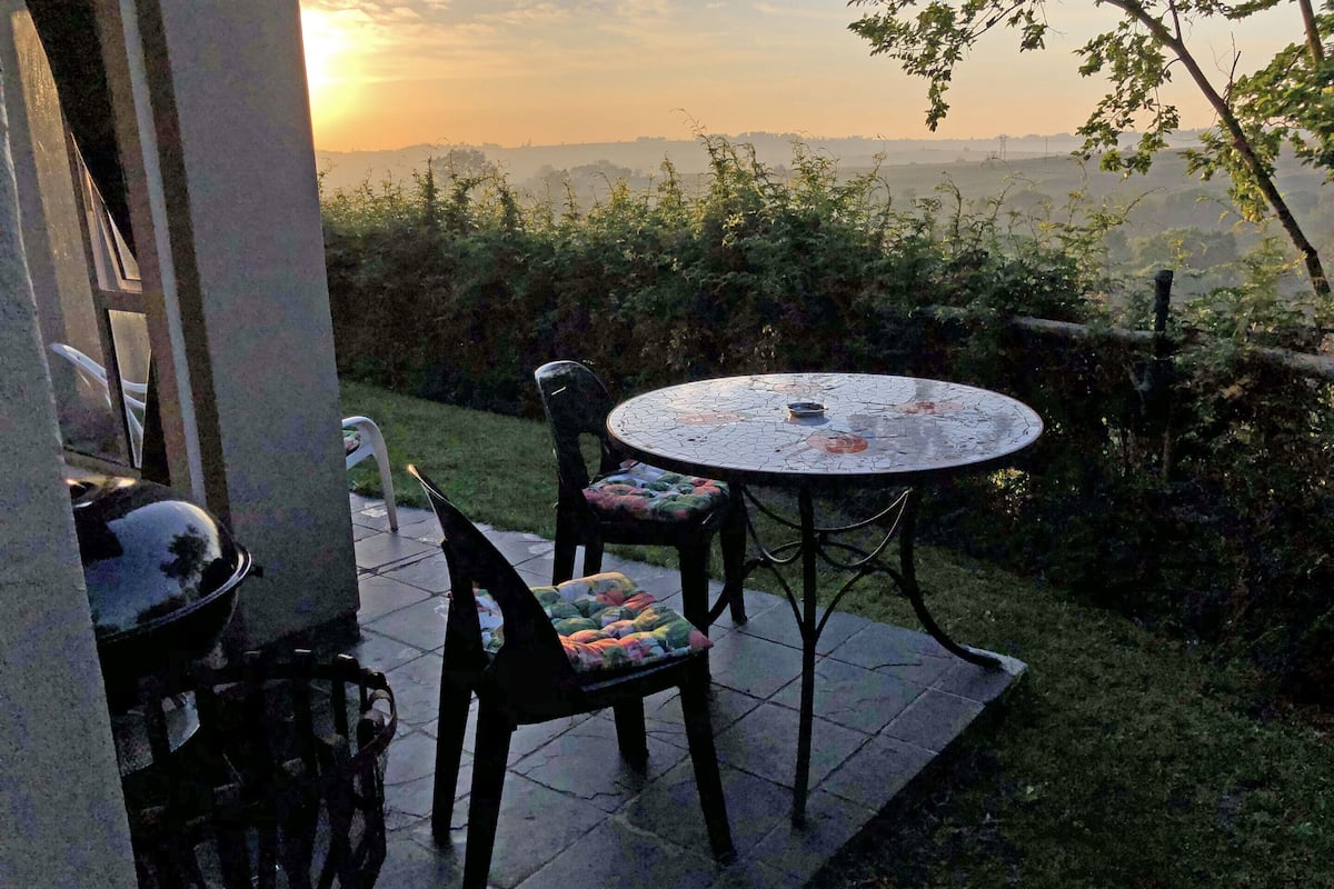 An outdoor patio with a round table and two chairs, surrounded by greenery and a view of the landscape.