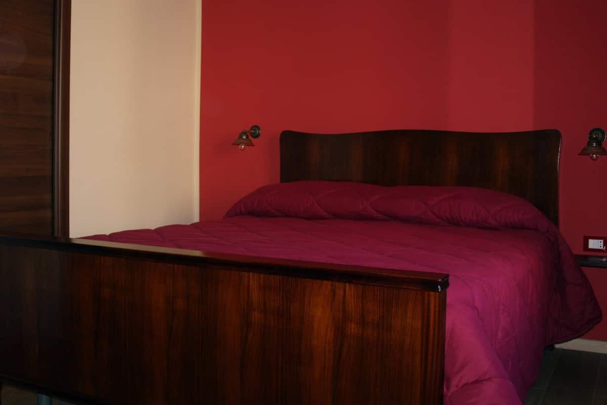 A bedroom with a wooden bed frame, a red wall, and two wall-mounted lamps.