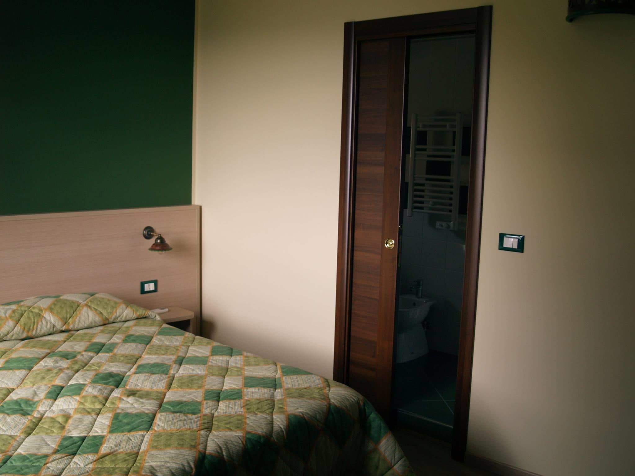 A bedroom with a wooden headboard, a quilted bedspread, a door leading to a bathroom, and a light switch on the wall.