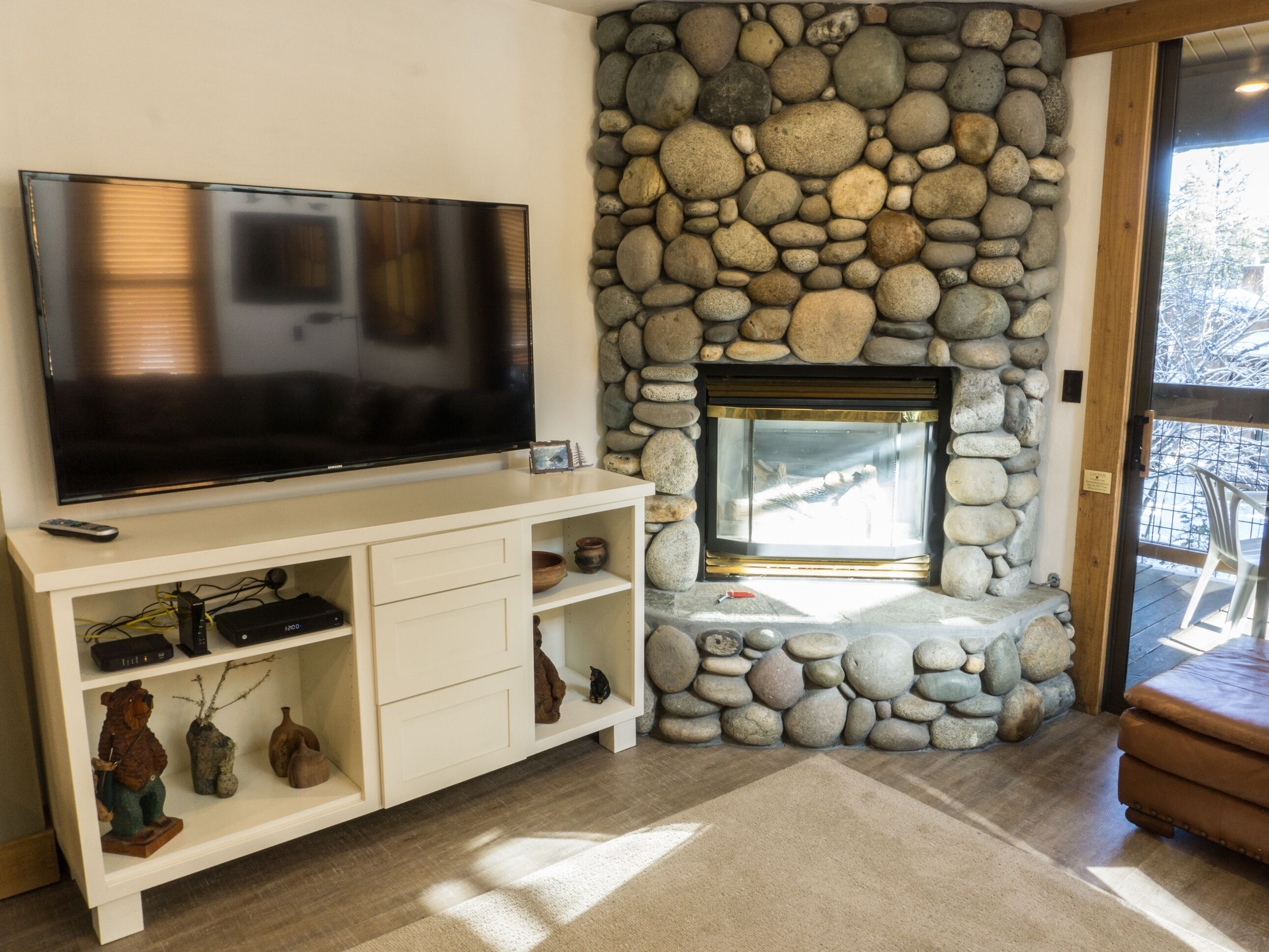 A living room with a stone fireplace, a flat-screen TV on a white entertainment unit, and a wooden bench.