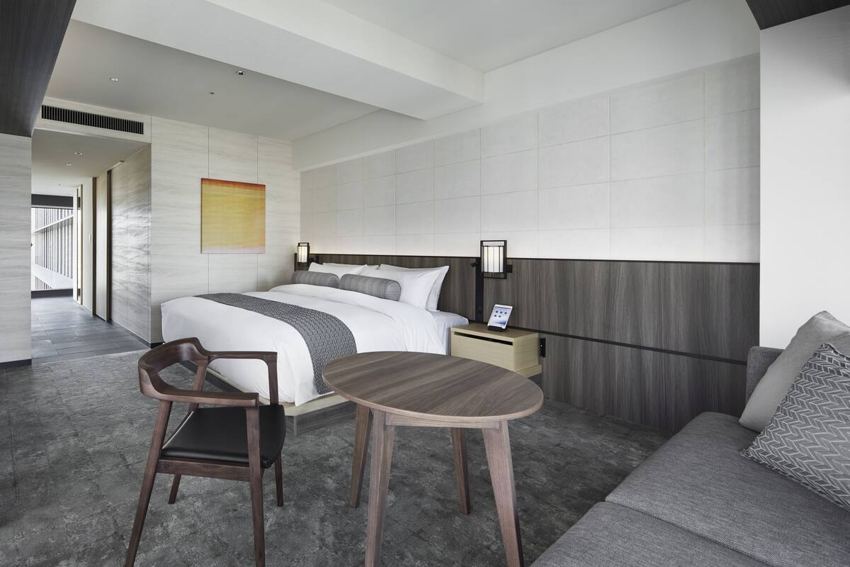 Una habitación de hotel moderna con una cama grande, una mesa de madera, un sofá y una mesita de noche con lámpara.