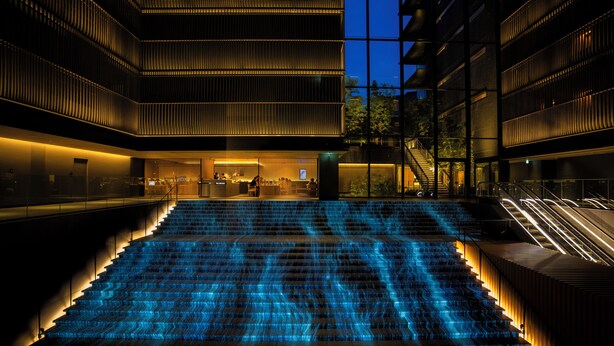 Een modern gebouw met een centrale trap, verlicht met blauw licht, omgeven door glazen balustrades en een zwembad.