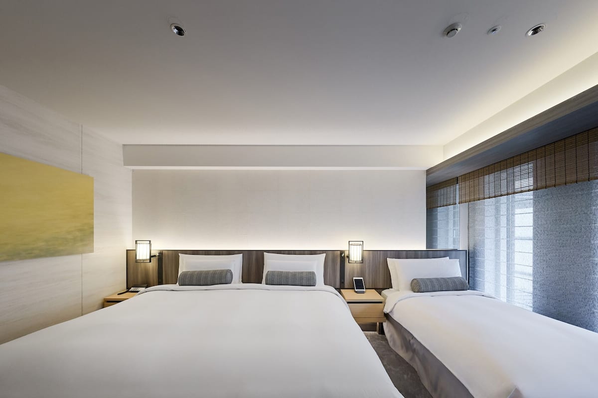 Habitación de hotel moderna con una cama grande, mesitas de noche y un amplio ventanal con persianas.