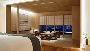 Een moderne hotelkamer met een houten vloer, een bed met beddengoed met een patroon, een eettafel met stoelen en uitzicht door grote shoji-schermen.