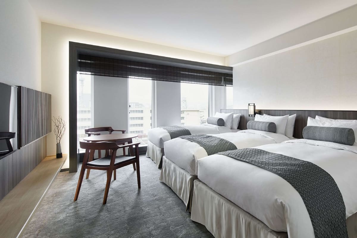 Una habitación de hotel moderna con una cama grande, un escritorio y una silla. Hay una ventana con persianas, un televisor de pantalla plana montado en la pared y un jarrón decorativo en el escritorio.