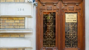 Entrada de hotel con puerta de madera, ventanal con reja decorativa y cartel con el nombre y la dirección del hotel.