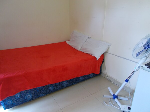 A small room with a single bed, a white wall, and a standing fan.