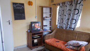 Un salon avec un téléviseur posé sur un meuble en bois, un canapé en osier recouvert d’une couverture orange, une bibliothèque et un tableau noir sur lequel on a écrit au mur.