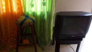Une pièce avec un téléviseur posé sur un meuble, un tabouret en bois avec un coussin à motifs, et un rideau aux motifs verts et oranges.