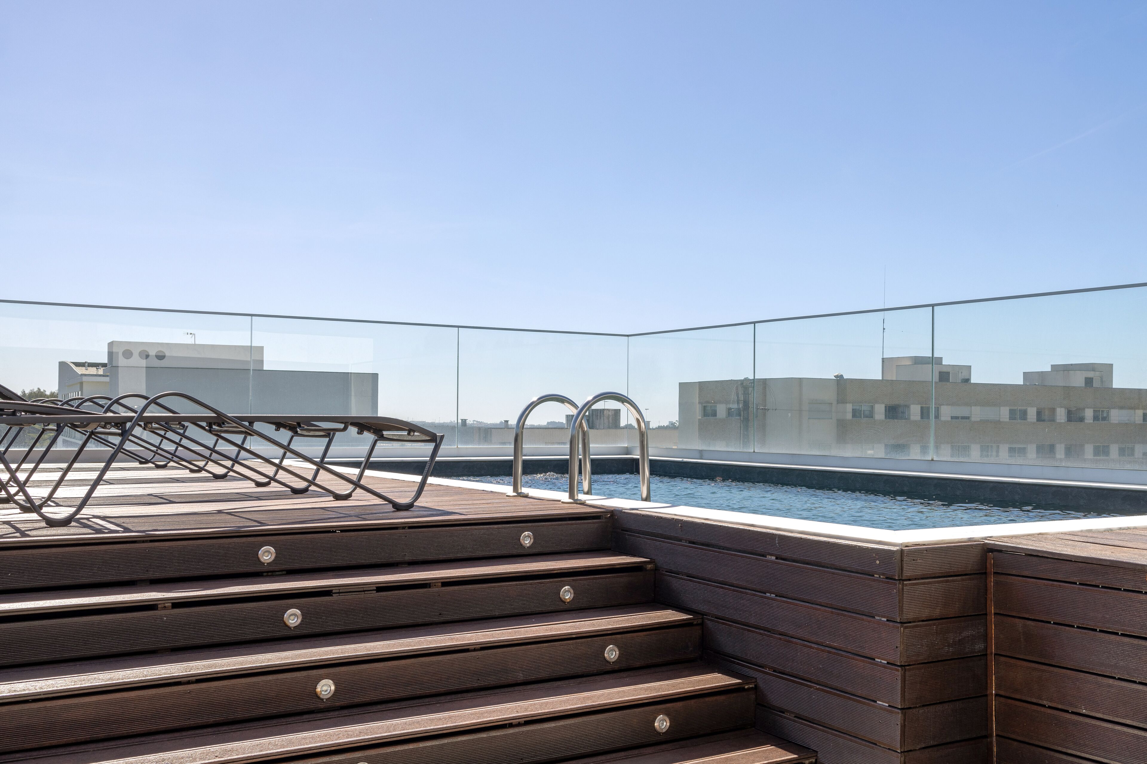 A rooftop pool area with wooden steps leading to the pool, metal railings, and a clear view of the cityscape in the background.