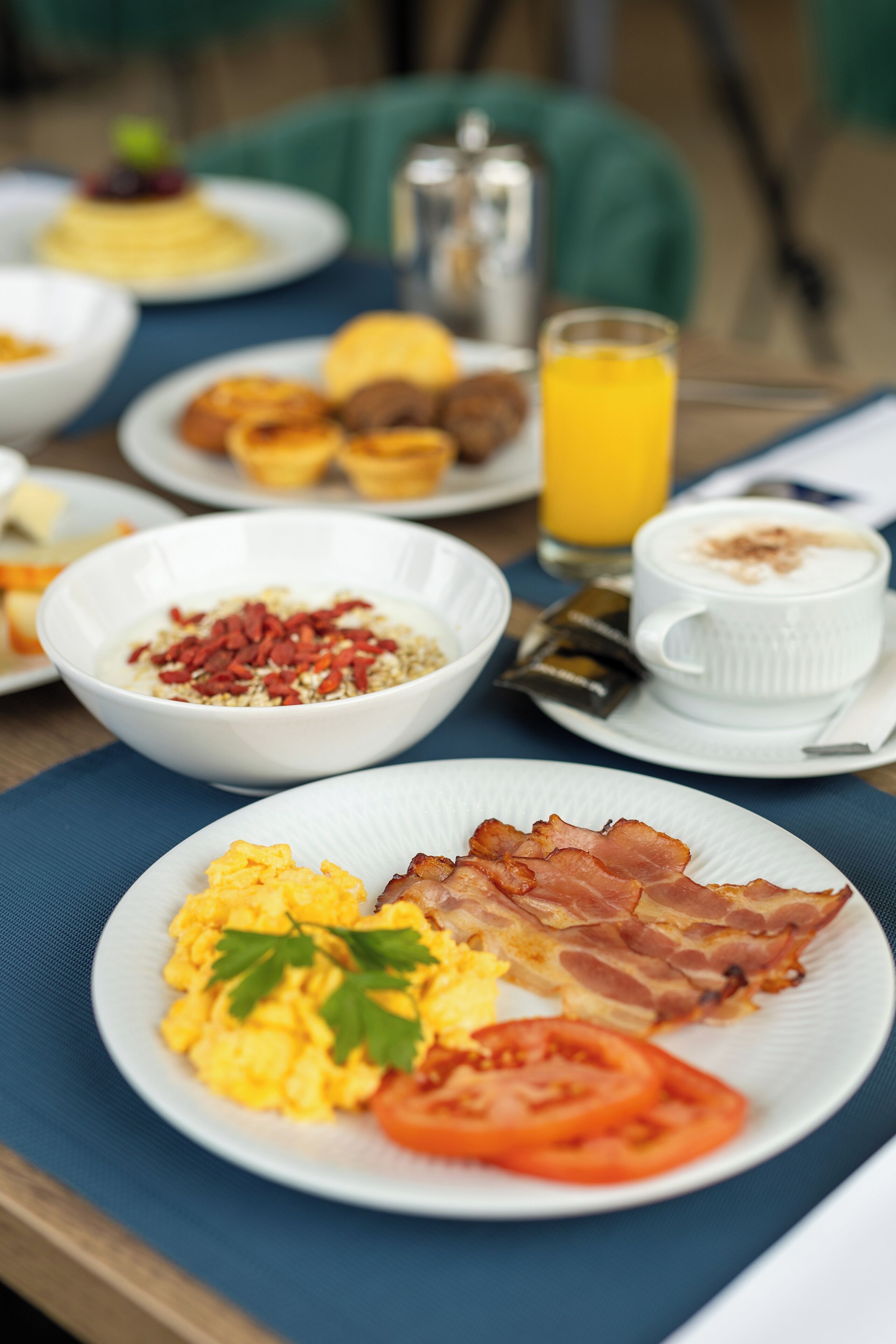 Una mesa de desayuno con huevos revueltos, tocino y tomates, un tazón de cereal y un vaso de jugo de naranja.