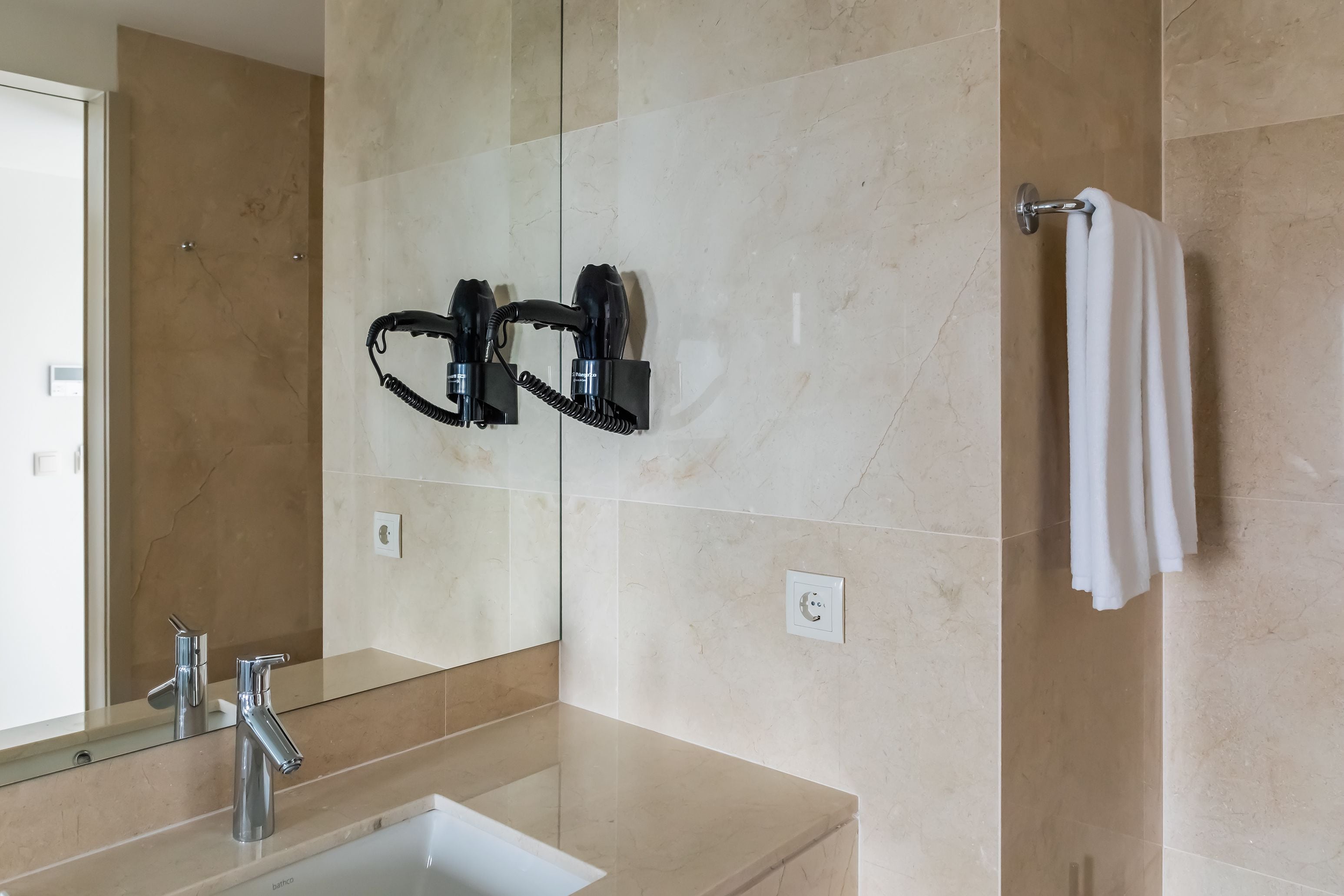 Une salle de bain avec un lavabo, deux sèche-cheveux noirs fixés au mur, une serviette suspendue à un porte-serviettes et un miroir.