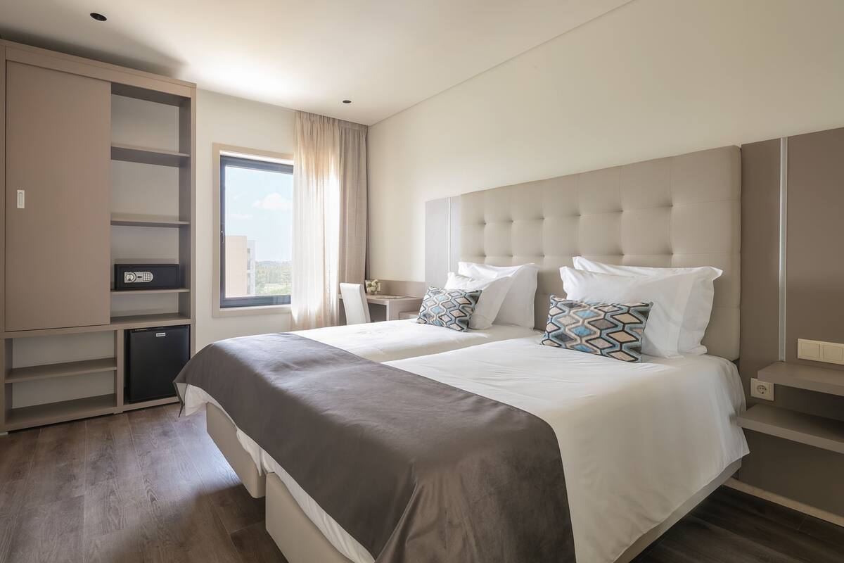 Moderni hotellihuone, jossa on kaksi sänkyä, sisäänrakennettu vaatekaappi, työpöytä ja näkymä kaupunkiin.