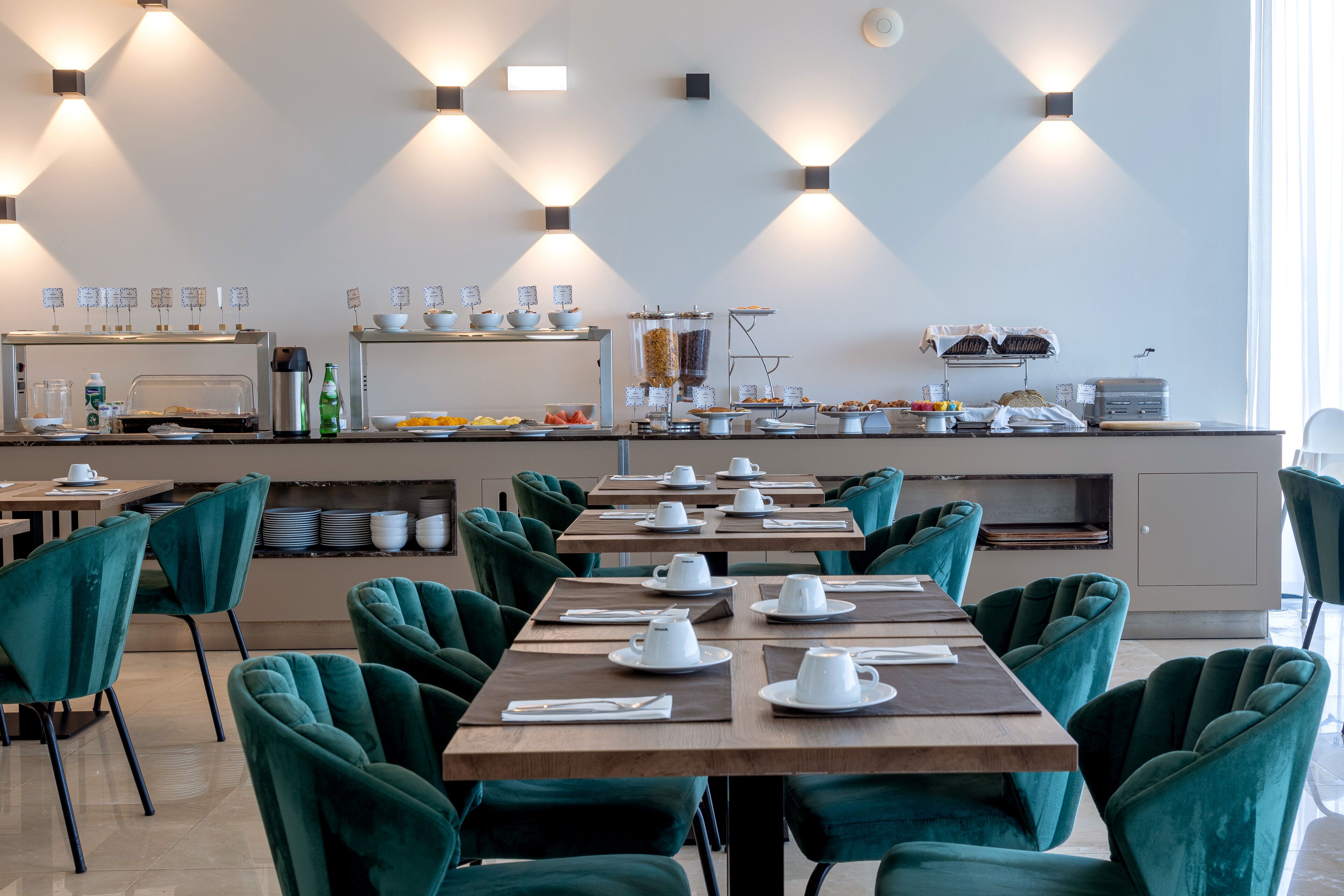 A modern dining area with teal chairs and tables, a buffet with various food items, and a coffee station.