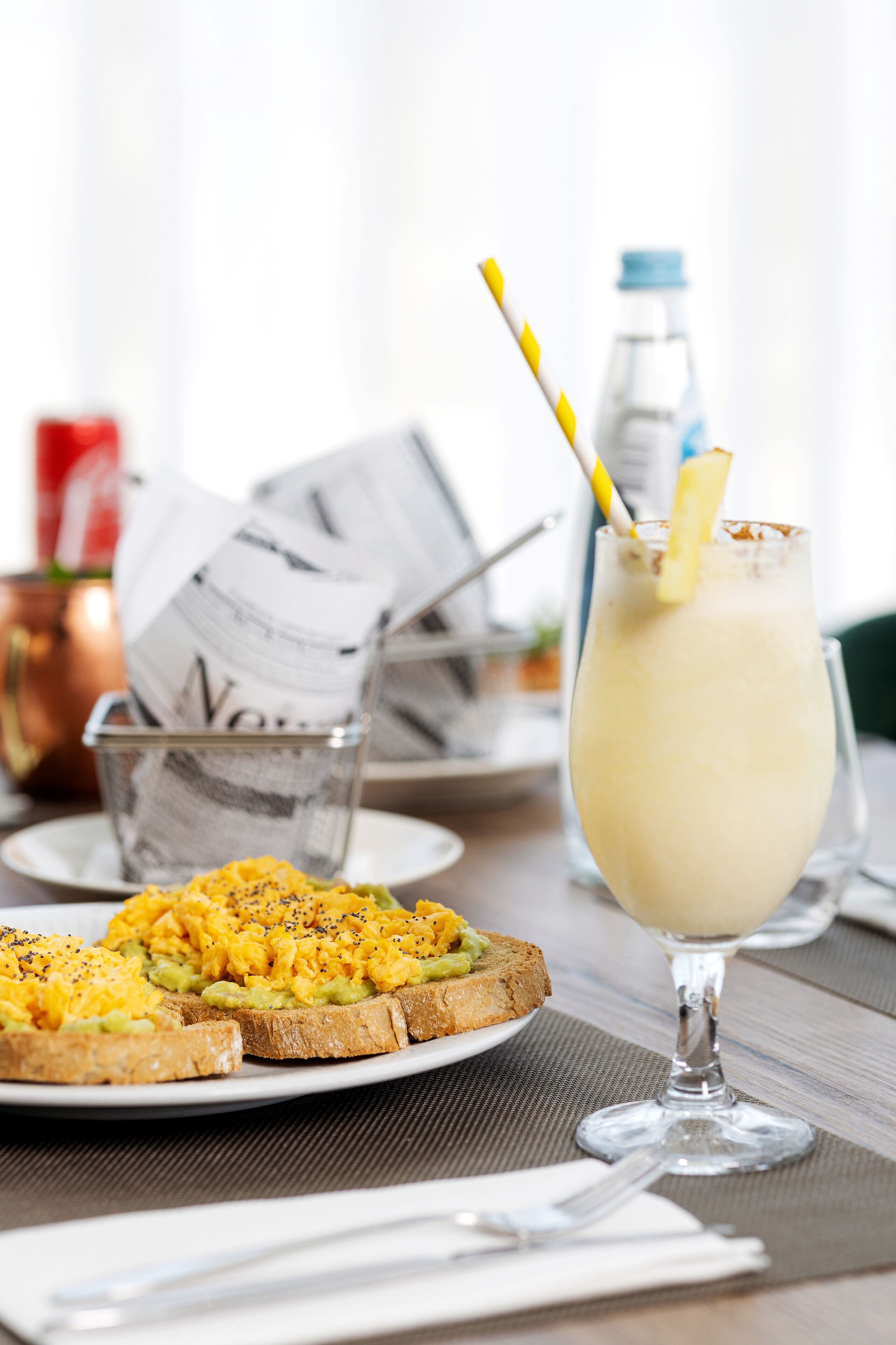 A breakfast setting with toast topped with scrambled eggs and avocado, a glass of a creamy beverage with a lemon slice and a striped straw, and a newspaper on a table.