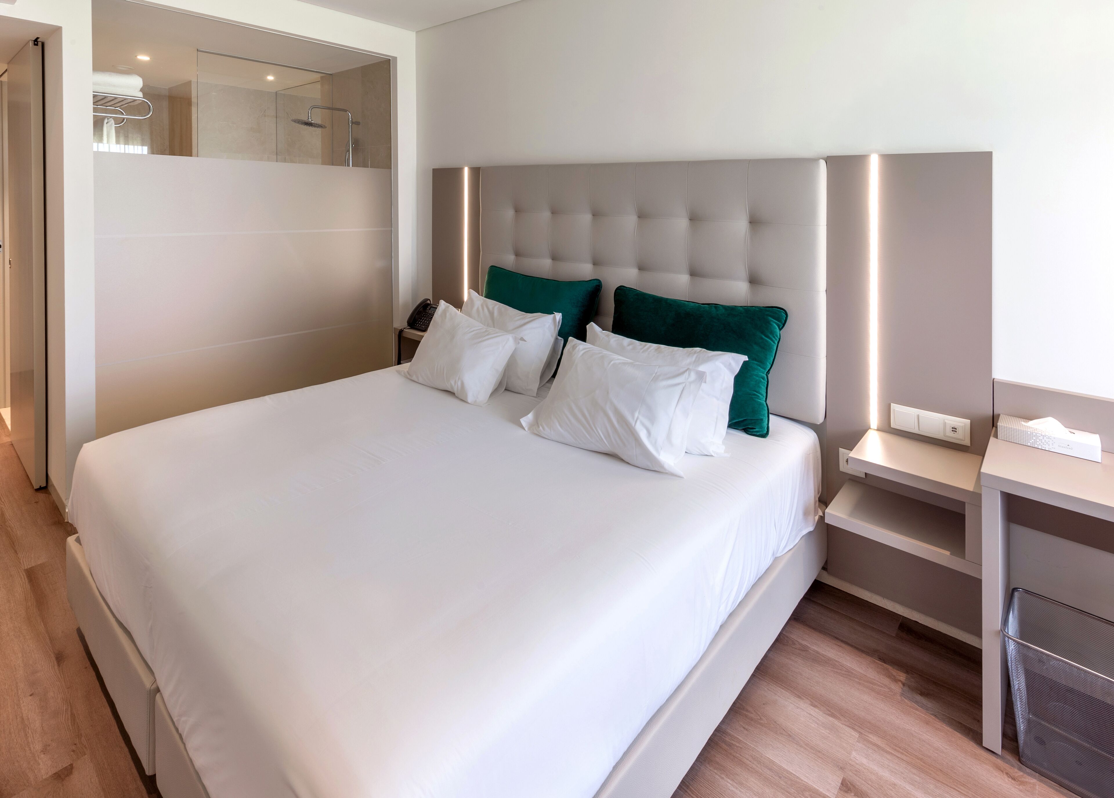 Ein modernes Hotelzimmer mit einem großen Bett, einem Nachttisch mit Lampe und einem durch eine Glastür sichtbaren Duschbereich.