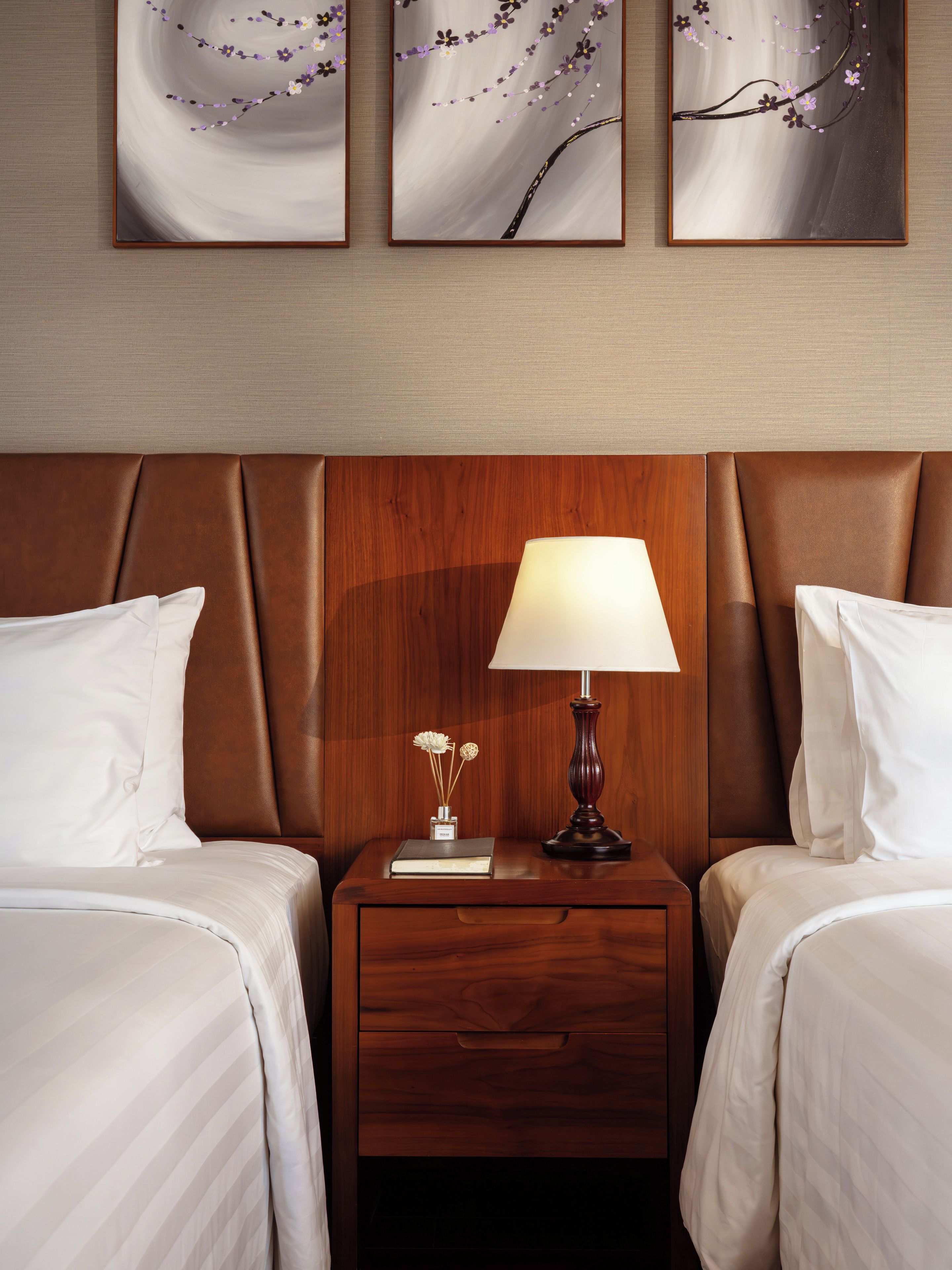 Quarto de hotel com duas camas, cabeceira de madeira, mesa de cabeceira com luminária e uma obra de arte na parede.