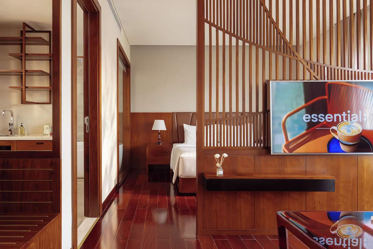 Een hotelkamer met een houten vloer, een bed met een wit hoofdeinde, een nachtkastje met een lamp, een aan de muur bevestigde televisie, en een houten trap.