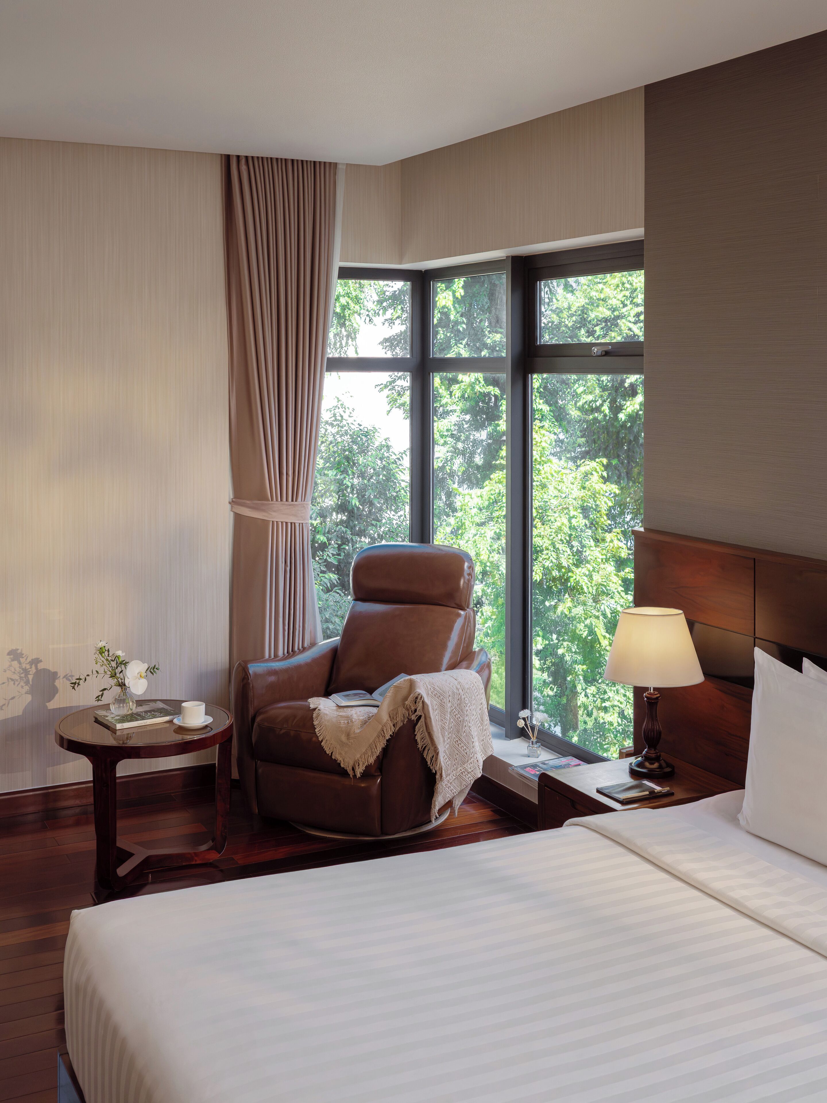 A hotel room with a large bed, a leather armchair, a small table with a lamp, and a view of greenery through the window.