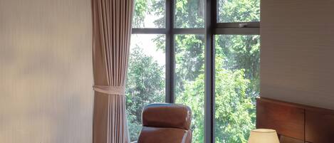A hotel room with a large bed, a leather armchair, a small table with a lamp, and a view of greenery through the window.