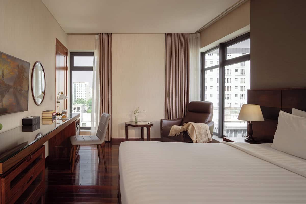 Hotelkamer met een bed, een bureau met spiegel, een stoel, een klein tafeltje met vaas en grote ramen met gordijnen.