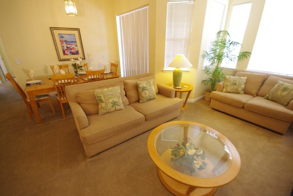 Una sala de estar con dos sofás color beige, una mesa de centro de cristal, una mesa de comedor con sillas y un cuadro en la pared.