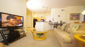 A living room with a TV, a sofa, a coffee table, and a kitchen in the background. - Ly53805 - Terrace Ridge - 4 Bed 2 Baths Condo (Davenport)