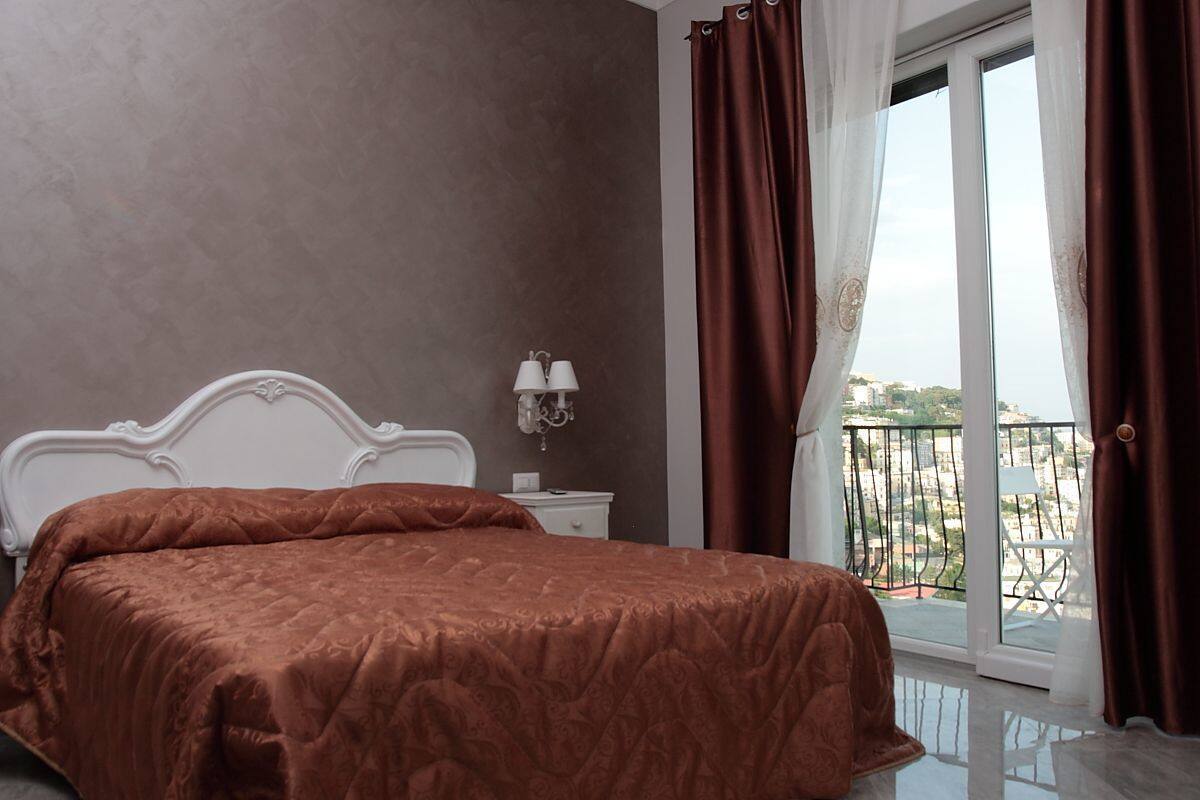 A bedroom with a large bed, a nightstand with lamps, a window with curtains, and a balcony view.