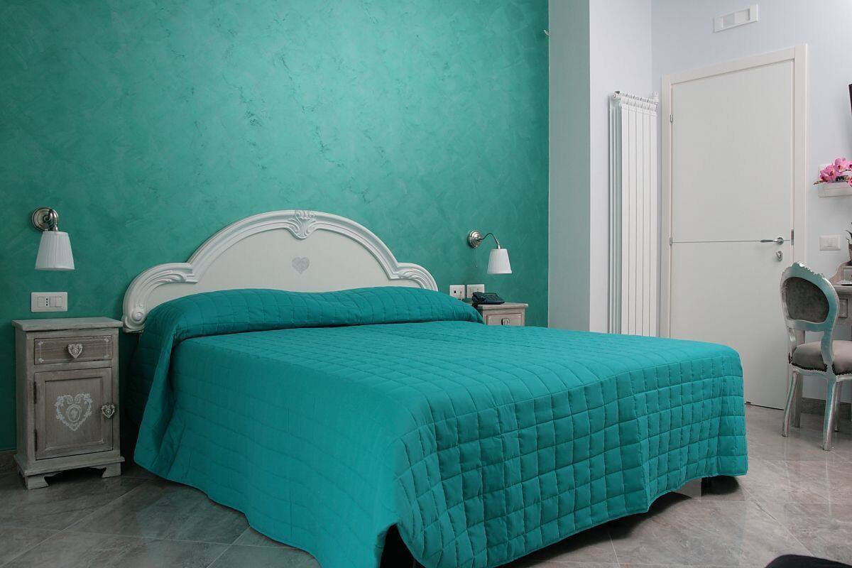 A bedroom with teal walls, a white bed with a quilted teal bedspread, a nightstand with a lamp, and a chair.