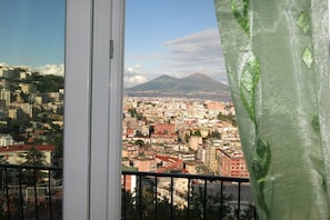 A cityscape with a mountain in the background, viewed from a balcony with a curtain partially covering the window. - Nido Dei Gabbiani (Naples)