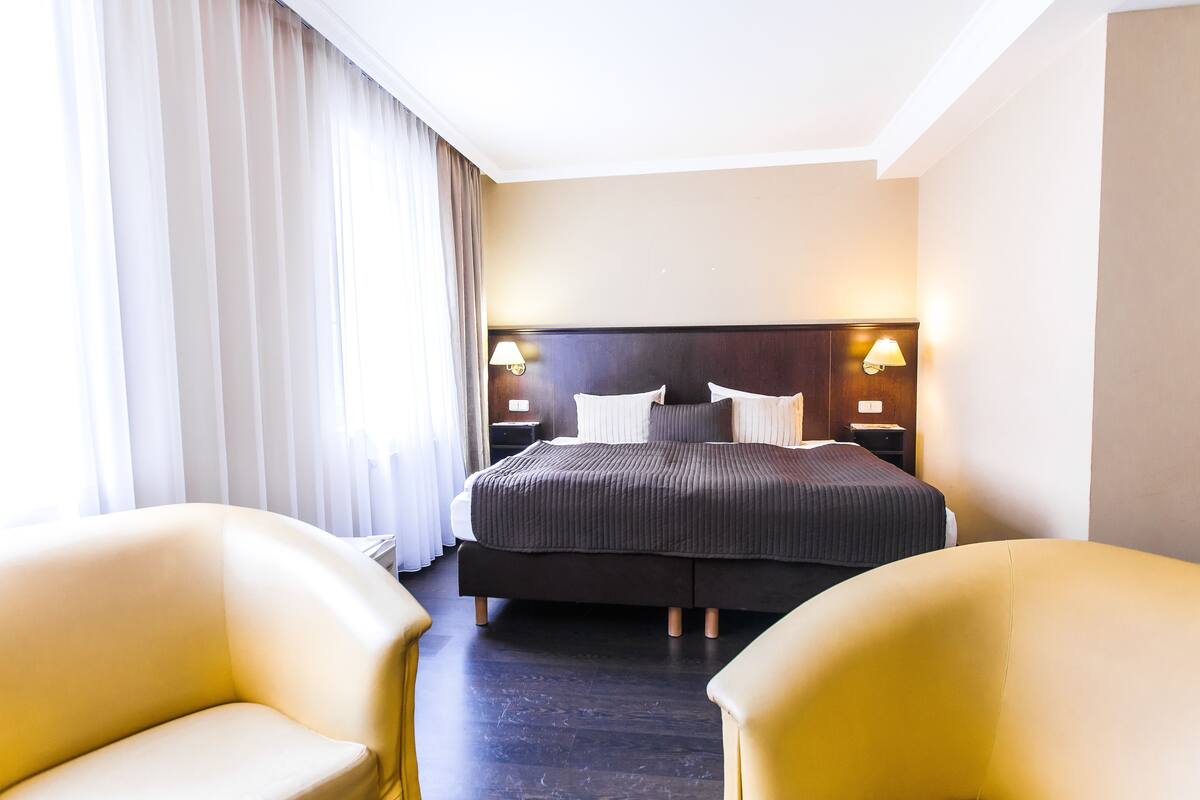 Een hotelkamer met een groot bed, twee nachtkastjes met lampen, een raam met gordijnen en twee gele fauteuils.