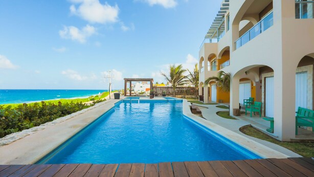 A swimming pool with a wooden deck in front, surrounded by a building with balconies and a view of the ocean. - Isla 33 - Studio 2413 (Isla Mujeres)