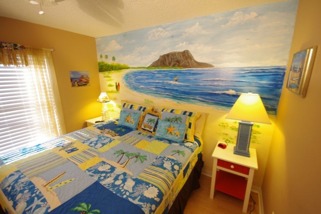 A bedroom with a beach-themed bedspread, a painting of a beach scene, and a nightstand with a lamp.