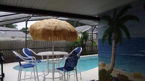A covered outdoor area with a pool, a table with chairs, and a mural of a beach scene.