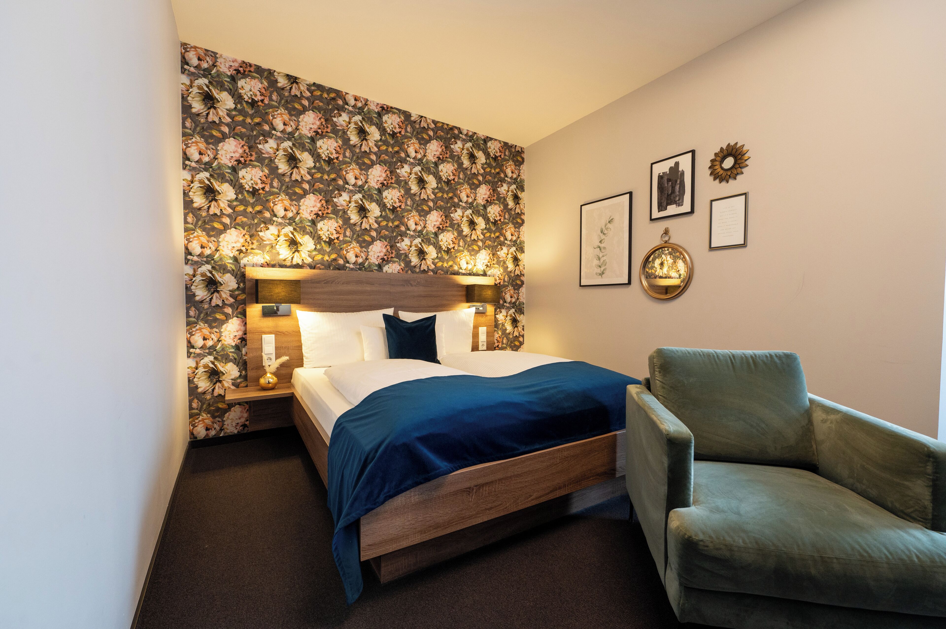 A neatly arranged bedroom with a floral wallpaper, a wooden headboard, a blue bedspread, and a green armchair.