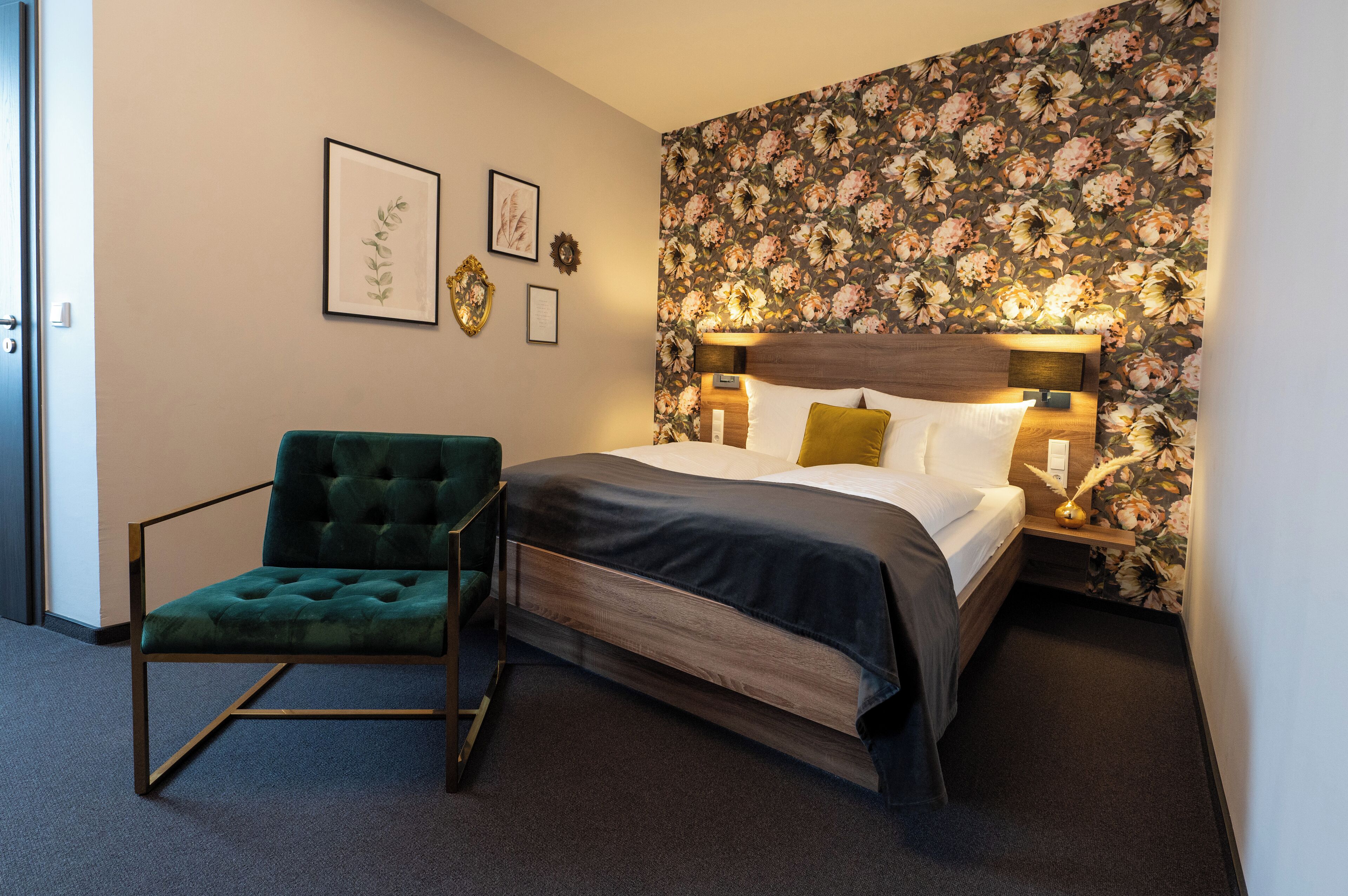 Una habitación de hotel con cama, un sillón verde, papel tapiz floral y obras de arte en la pared.