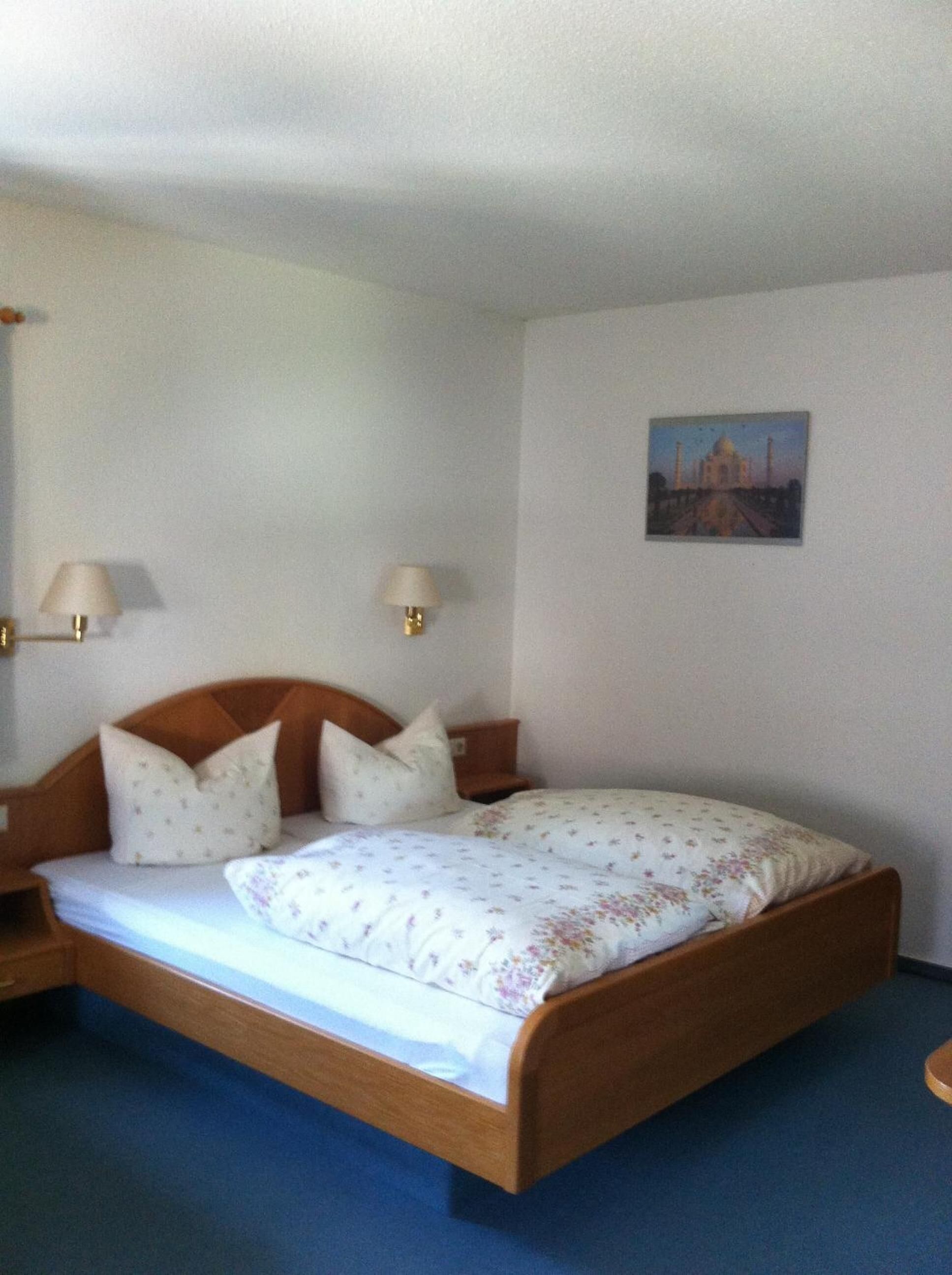 Un lit simple avec une tête de lit en bois, deux lampes de chevet, une couvre-lit à motifs floraux et un tableau représentant un bâtiment accroché au mur.