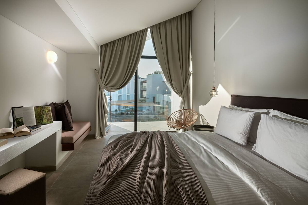 A modern hotel room with a large bed, a desk with a laptop, and a view of the cityscape through the window.
