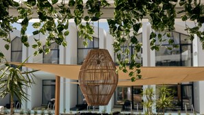 An outdoor seating area with a woven shade structure, potted plants, and a modern building in the background. - Cavo Vezal - Adults Only (Zakynthos)