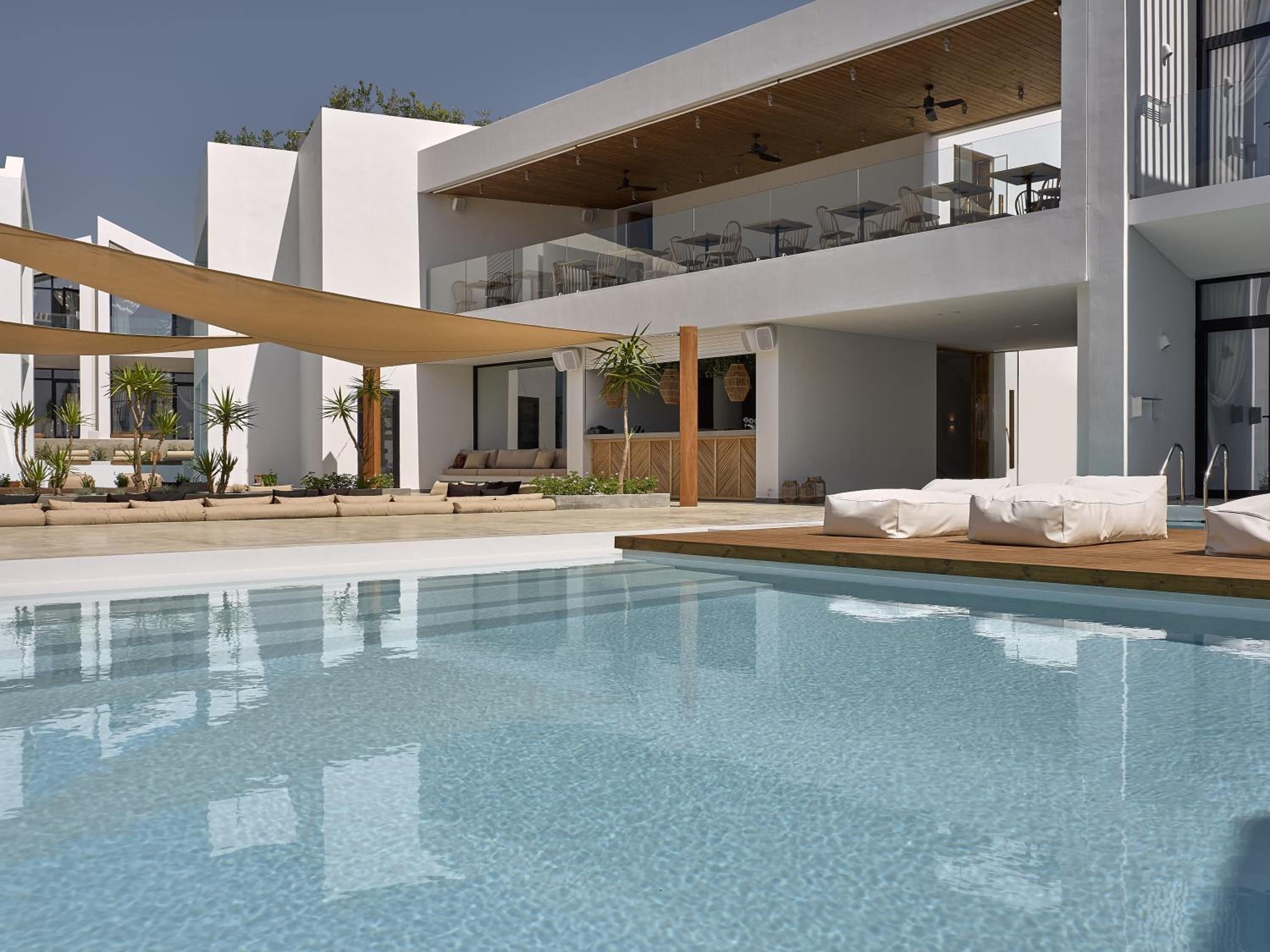 Un área de piscina moderna con sillones de descanso, una terraza de madera y un edificio de dos pisos con barandas de vidrio.