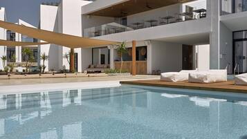 Un área de piscina moderna con sillones de descanso, una terraza de madera y un edificio de dos pisos con barandas de vidrio.