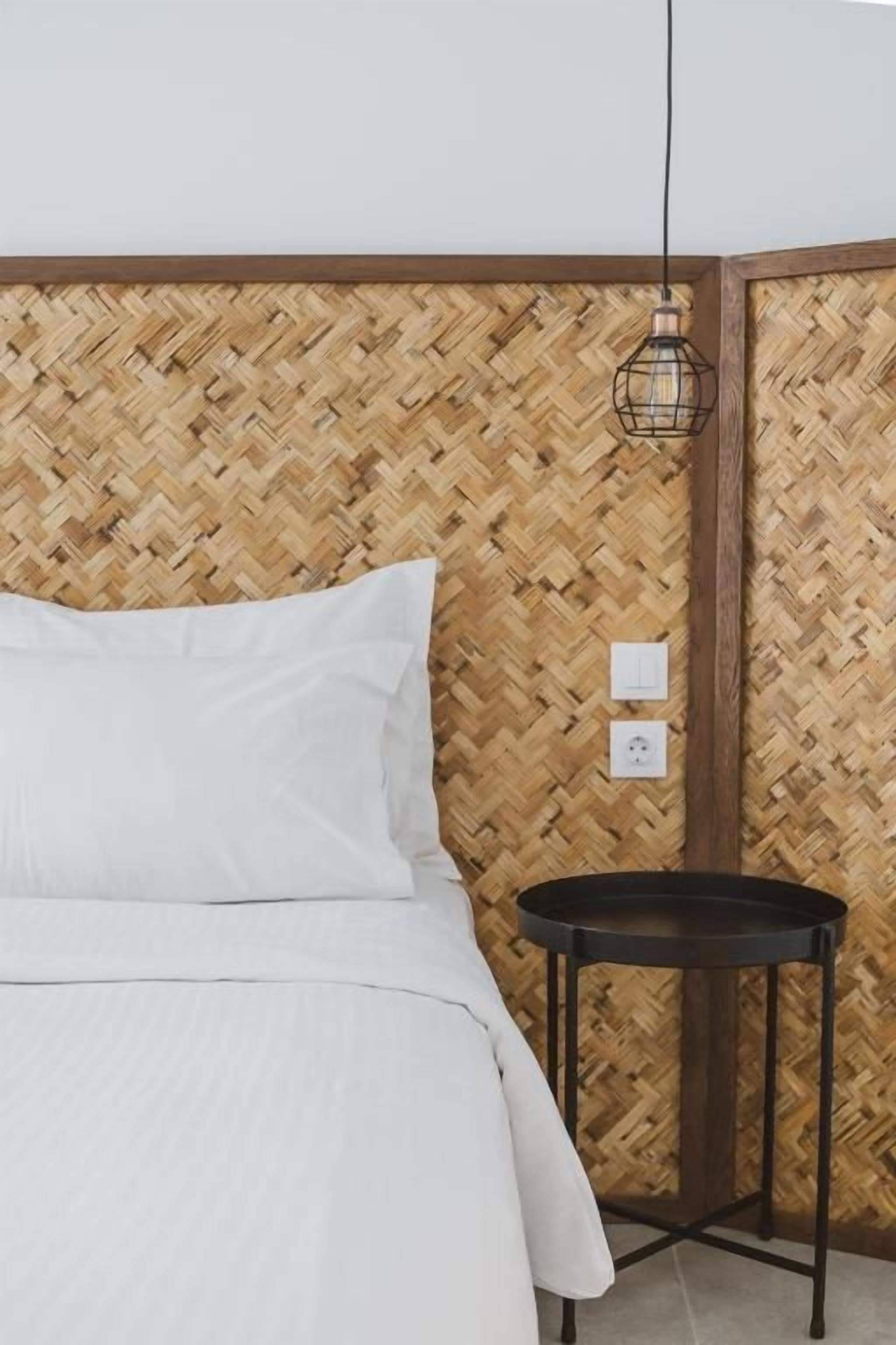 Una cama con ropa de cama blanca, una mesita de noche redonda negra y un panel decorativo tejido en la pared.