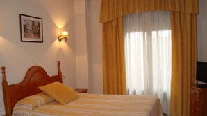 Una cama bien hecha con una colcha a rayas, un cabecero de madera y una sola almohada amarilla. Se ve una mesita de noche con una lámpara, un cuadro enmarcado en la pared y un televisor sobre un mueble de madera.