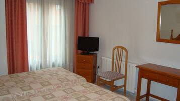 Une chambre à coucher avec un lit, une chaise en bois, un téléviseur posé sur une commode en bois et un miroir accroché au mur.