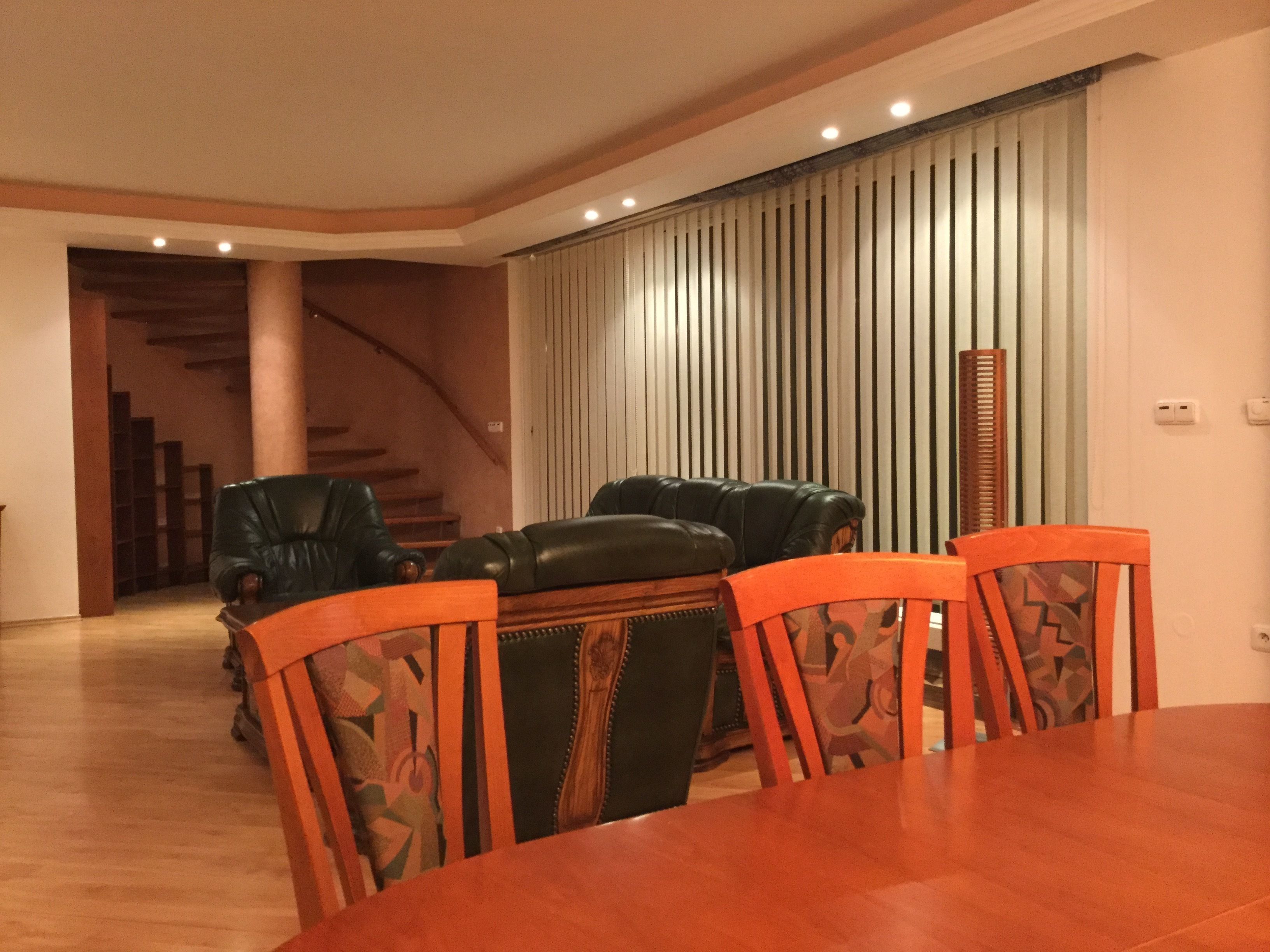 A dining area with a wooden table and chairs, a staircase in the background, and a sliding door with vertical blinds.