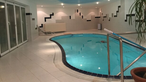 An indoor swimming pool with a curved design, tiled walls, and a ceiling with recessed lighting. - Justyna (Pardubice)