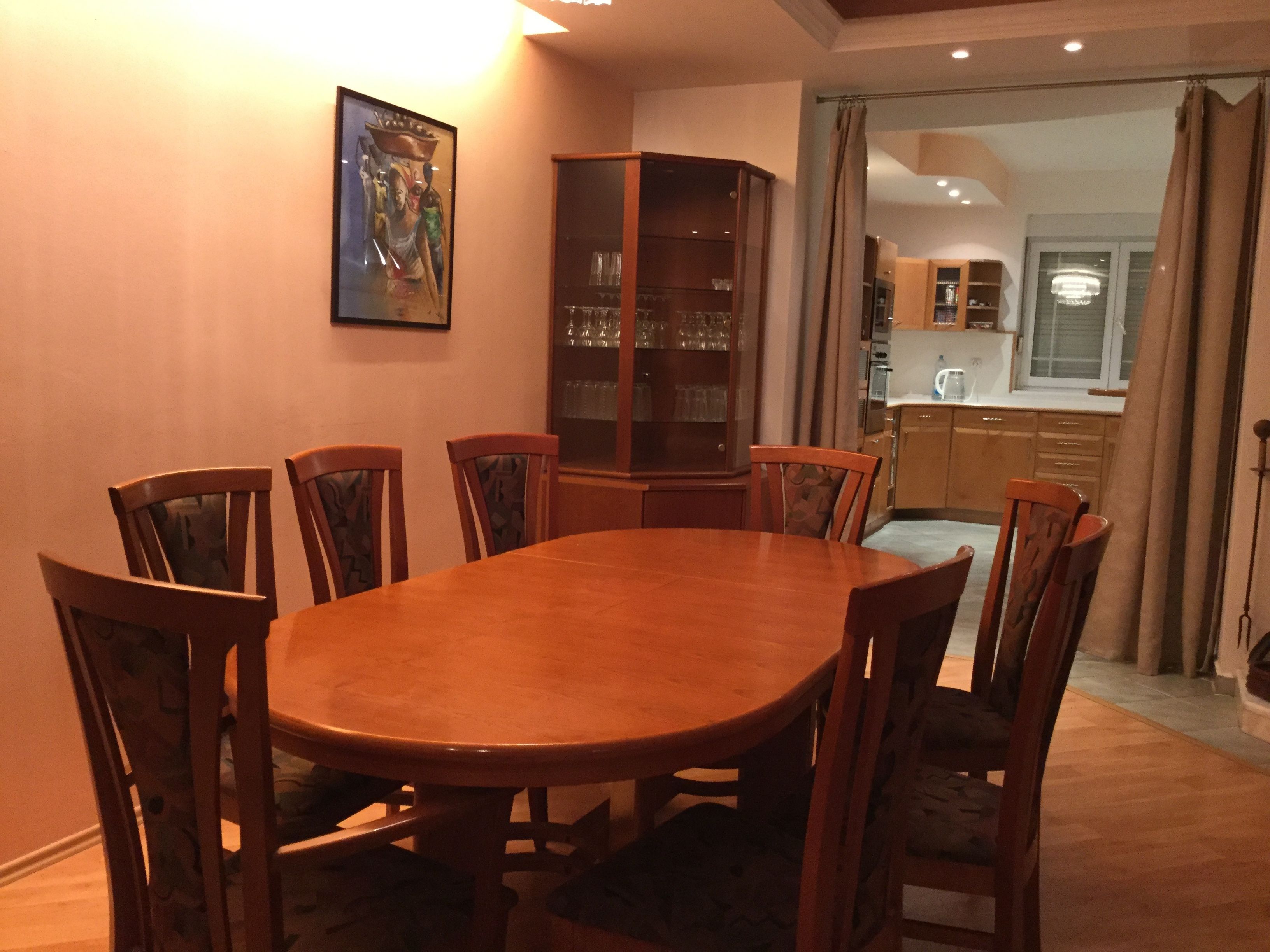 A dining area with a wooden table and chairs, a glass cabinet with wine glasses, and a painting on the wall.