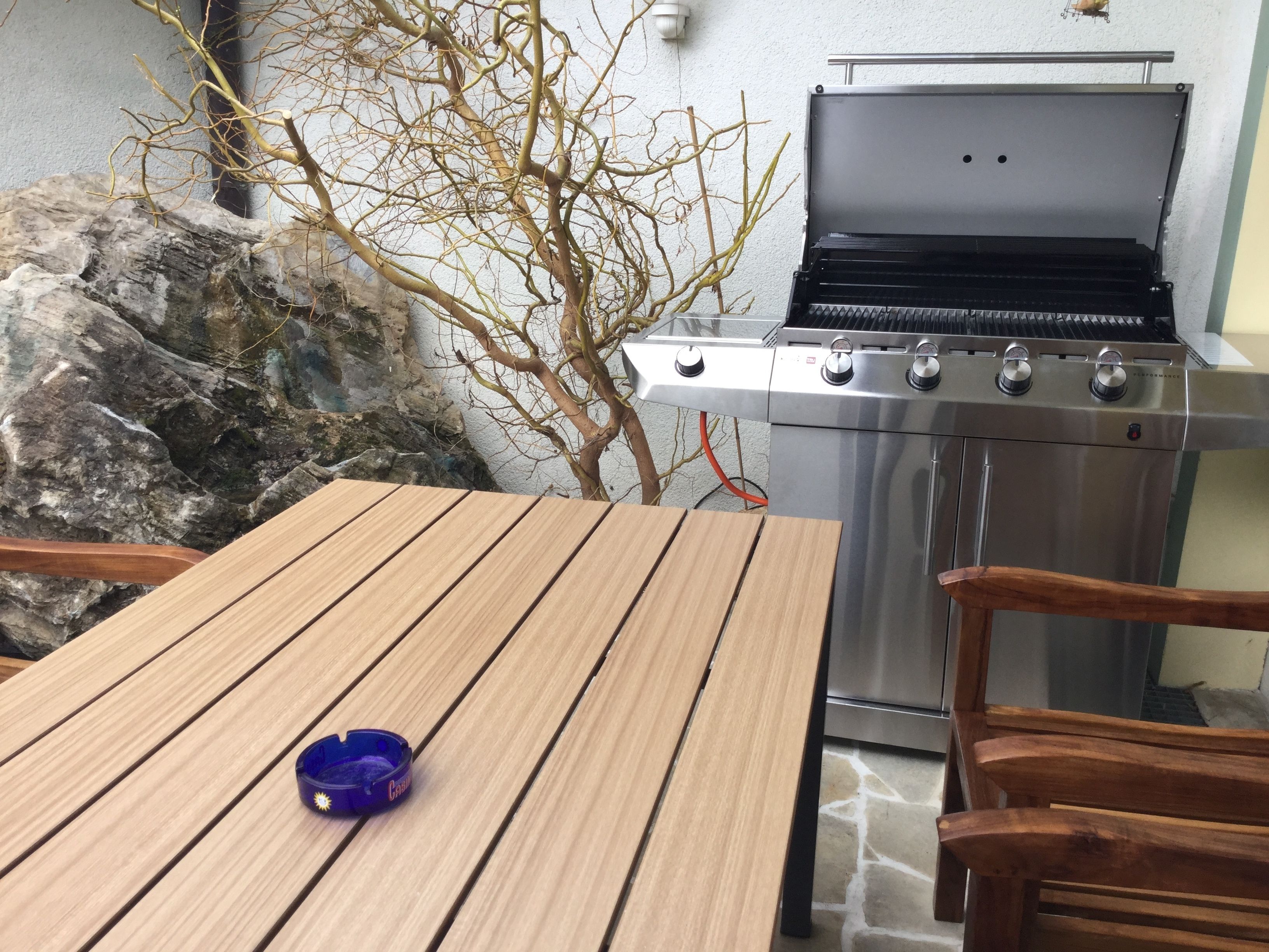 A wooden table with a blue ashtray, a stainless steel grill, and a stone wall with a bare tree branch.