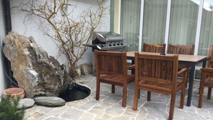 A patio area with stone flooring, a barbecue grill, wooden chairs, and a small rock garden. - Justyna (Pardubice)