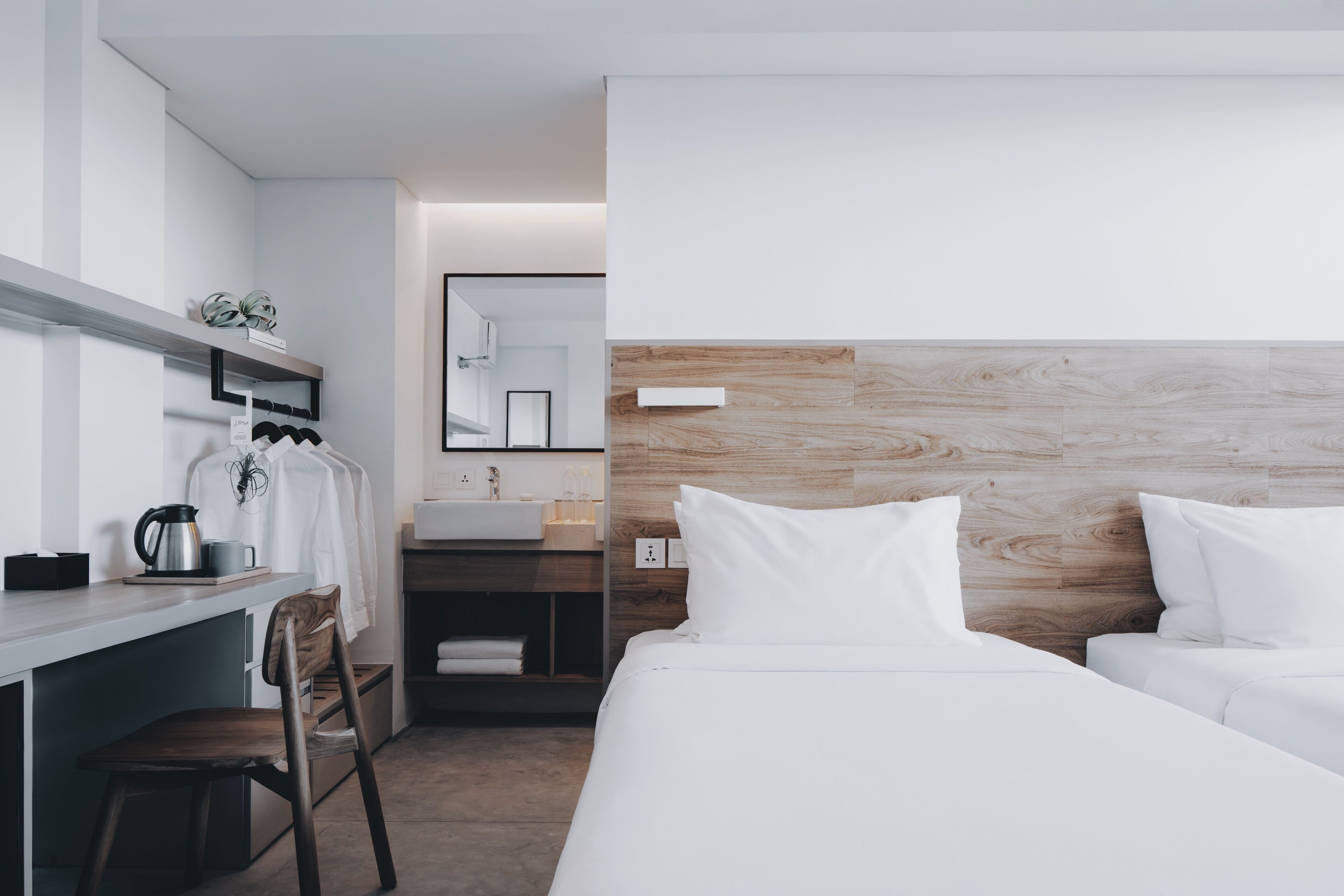 A modern hotel room with a wooden headboard, a white bed, a desk with a chair, and a small kitchenette.